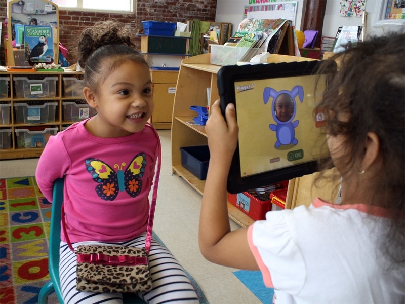 A preschooler taking a photo of another classmate using the iPad game Photo Friends.